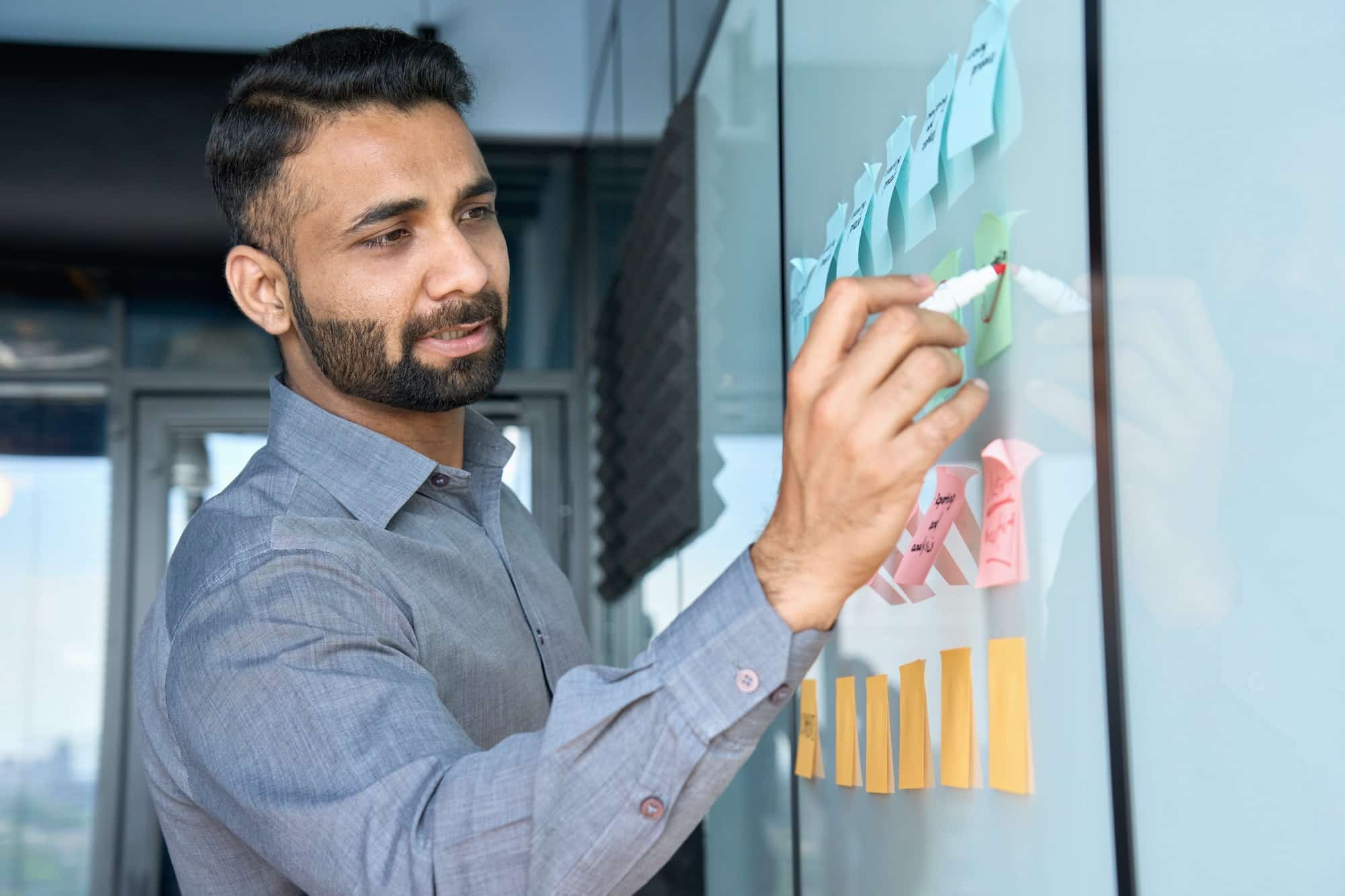 Young indian businessman standing writing on sticky notes on whiteboard.